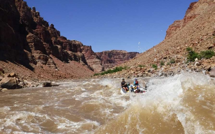People paddle a raft through whitewater framed by tall canyon walls. 